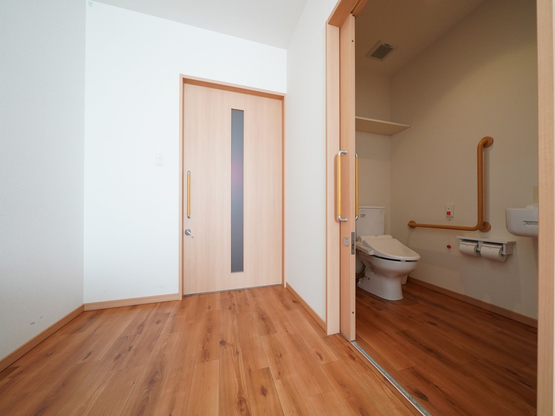 居室入口付近の幅の広い引き戸を開けるとトイレが設置されている。便座横の壁には大きな手すりが設置されている。