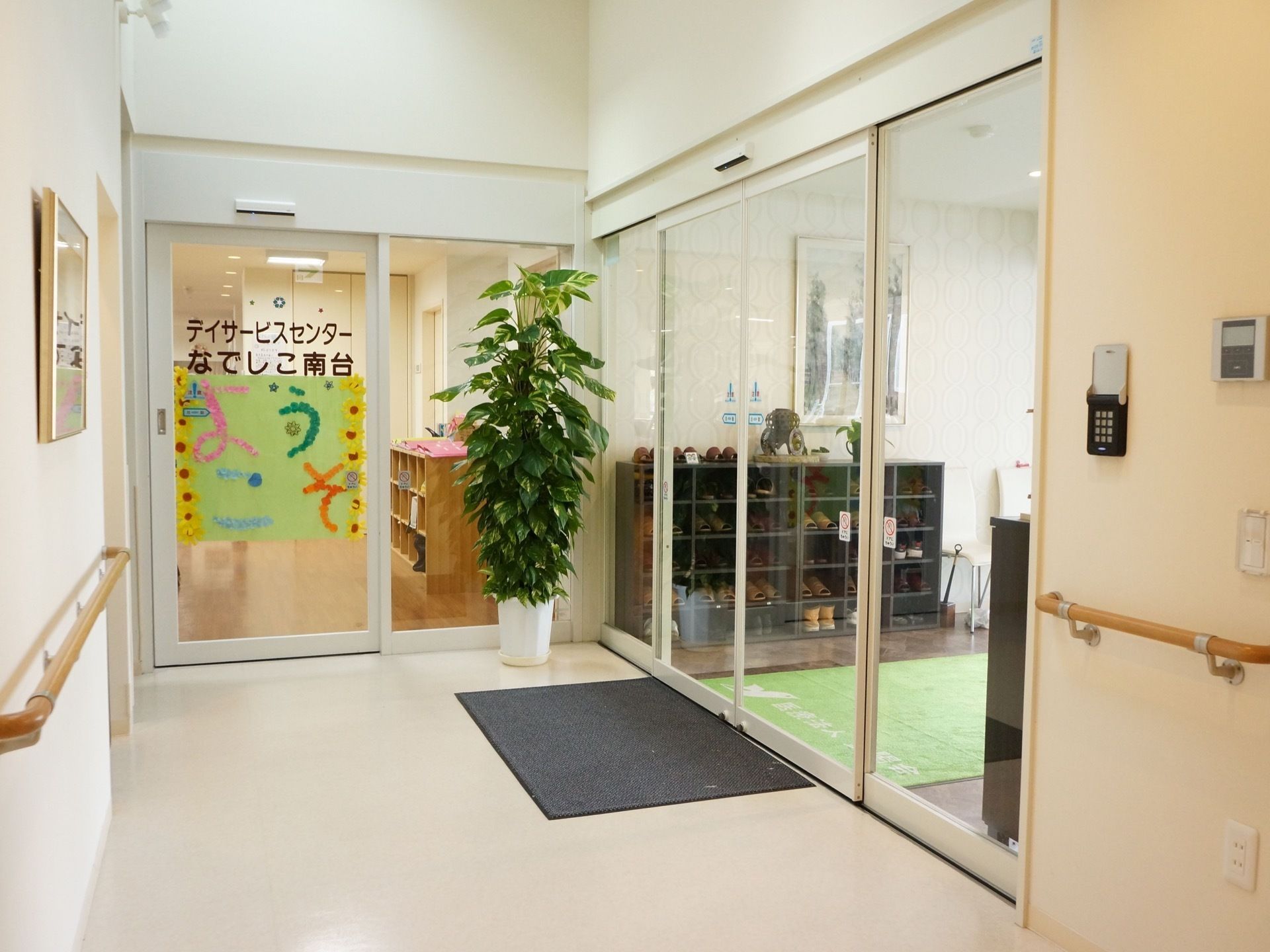 玄関を入ると右手にはデイサービスの自動ドアがあり、大きな観葉植物が置かれている。正面には絵が飾られている。