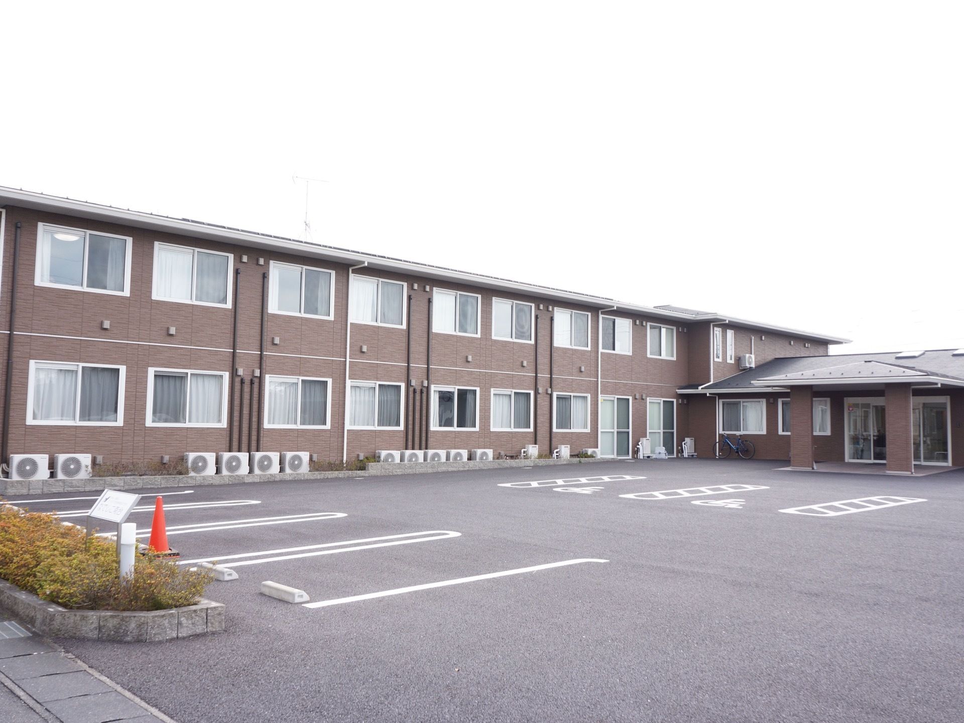 建物は2階建てになっており、落ち着いたブラウンの外壁になっている。施設の前に広い駐車スペースがある。