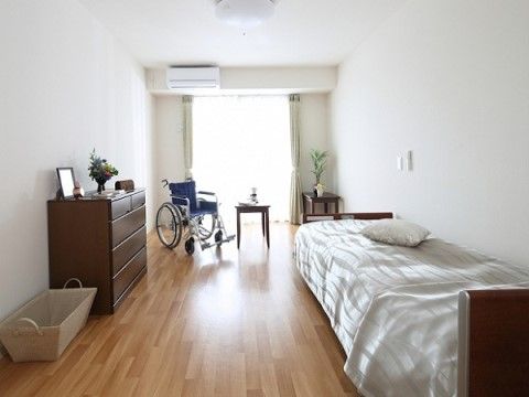 フローリングの床に白い壁の明るい部屋である。ベッドと車椅子と茶色いチェストが置いてあり、壁にエアコンが設置されている。