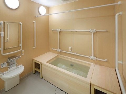 たくさんの手すりが取り付けられた、ベージュを基調とした広々とした浴室である。浴槽と壁の間のスペースに足のついた板がはめられている。