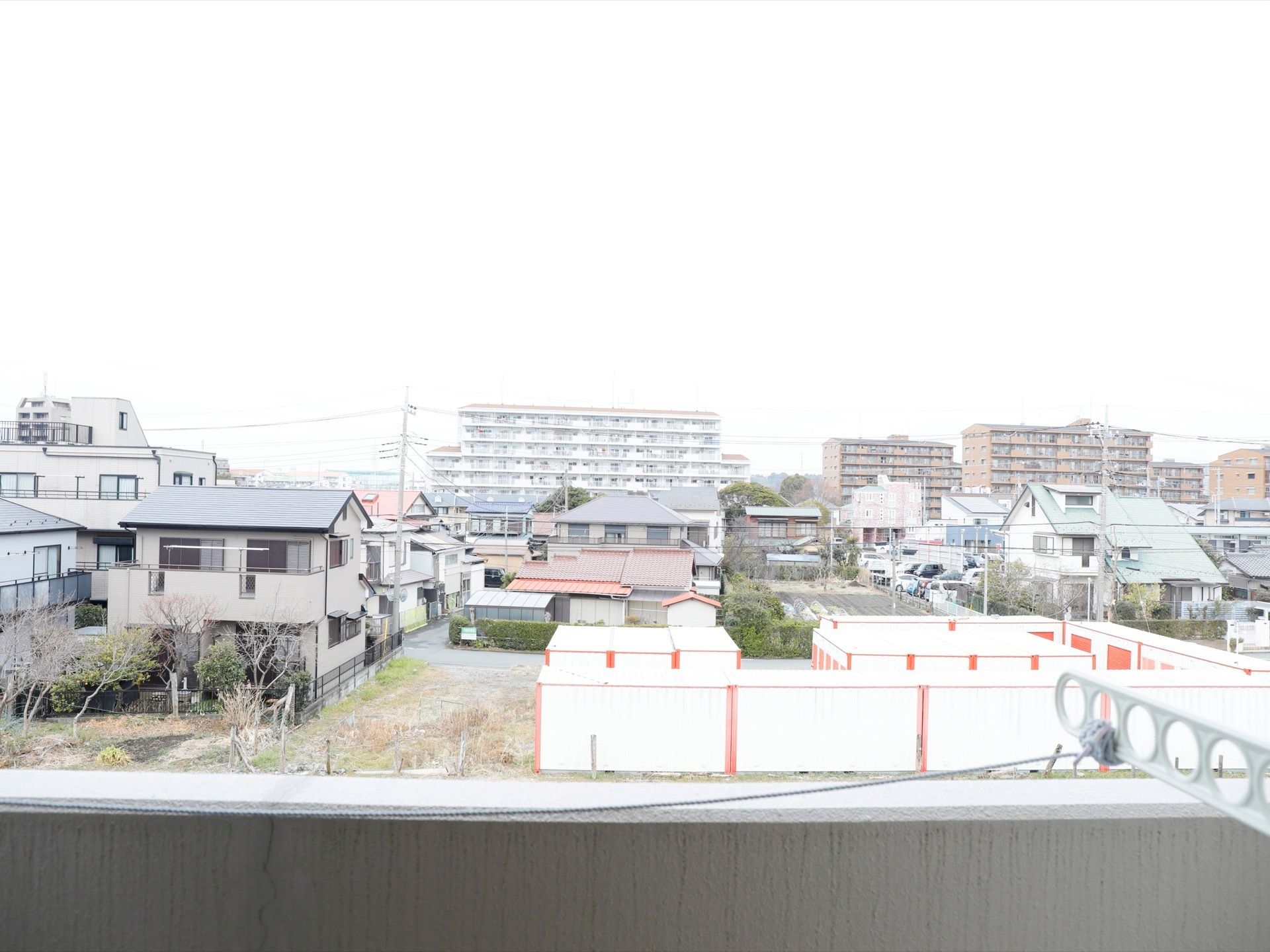 ベランダから見た景色。ベランダには物干しざお掛けが付いている。周囲は住宅街で沢山の家やマンションが見える。