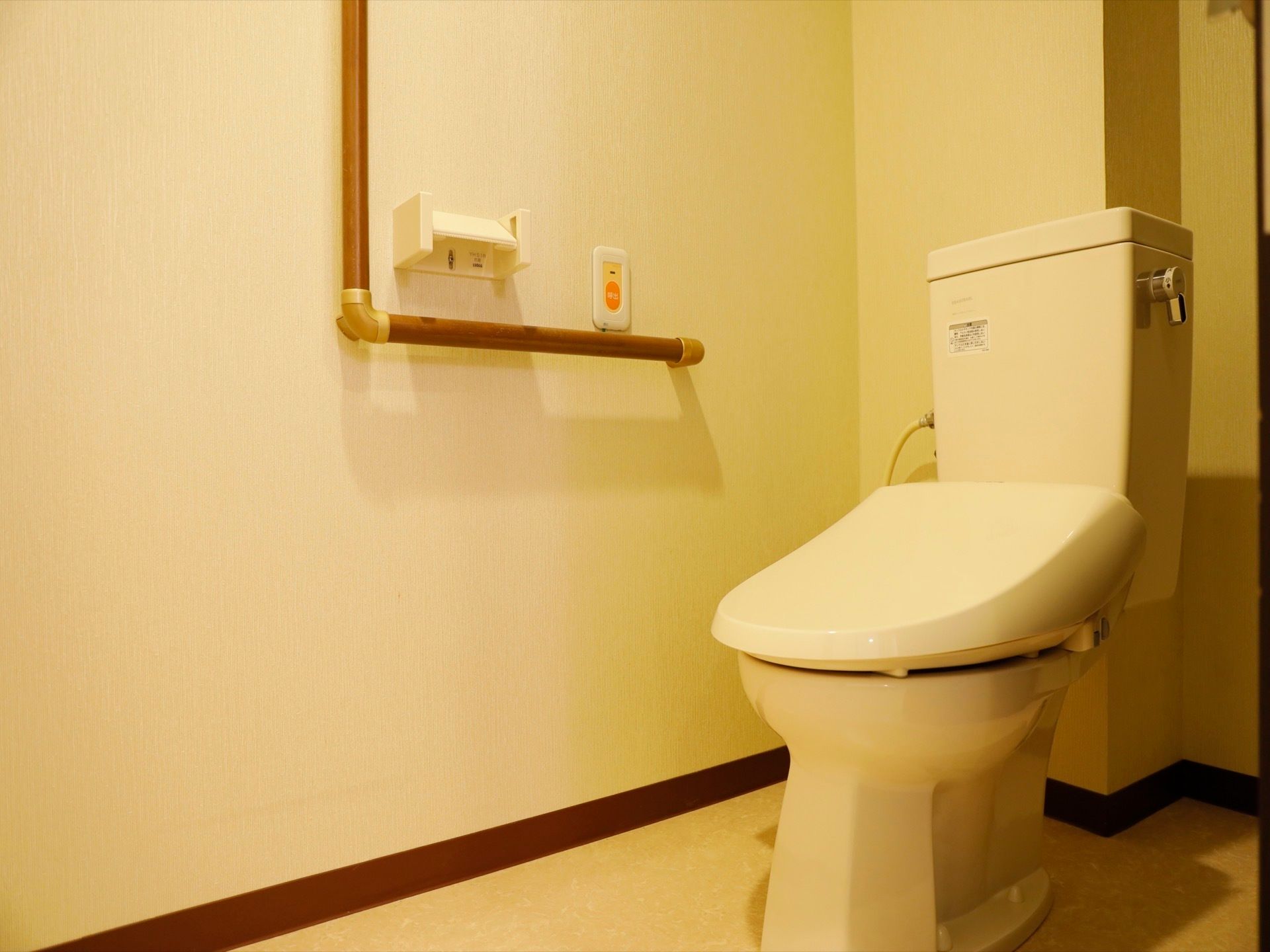 タンク付きの洋式トイレが設置されている。壁にはトイレットペーパーホルダー、L字の手すり、ナースコールが付いている。