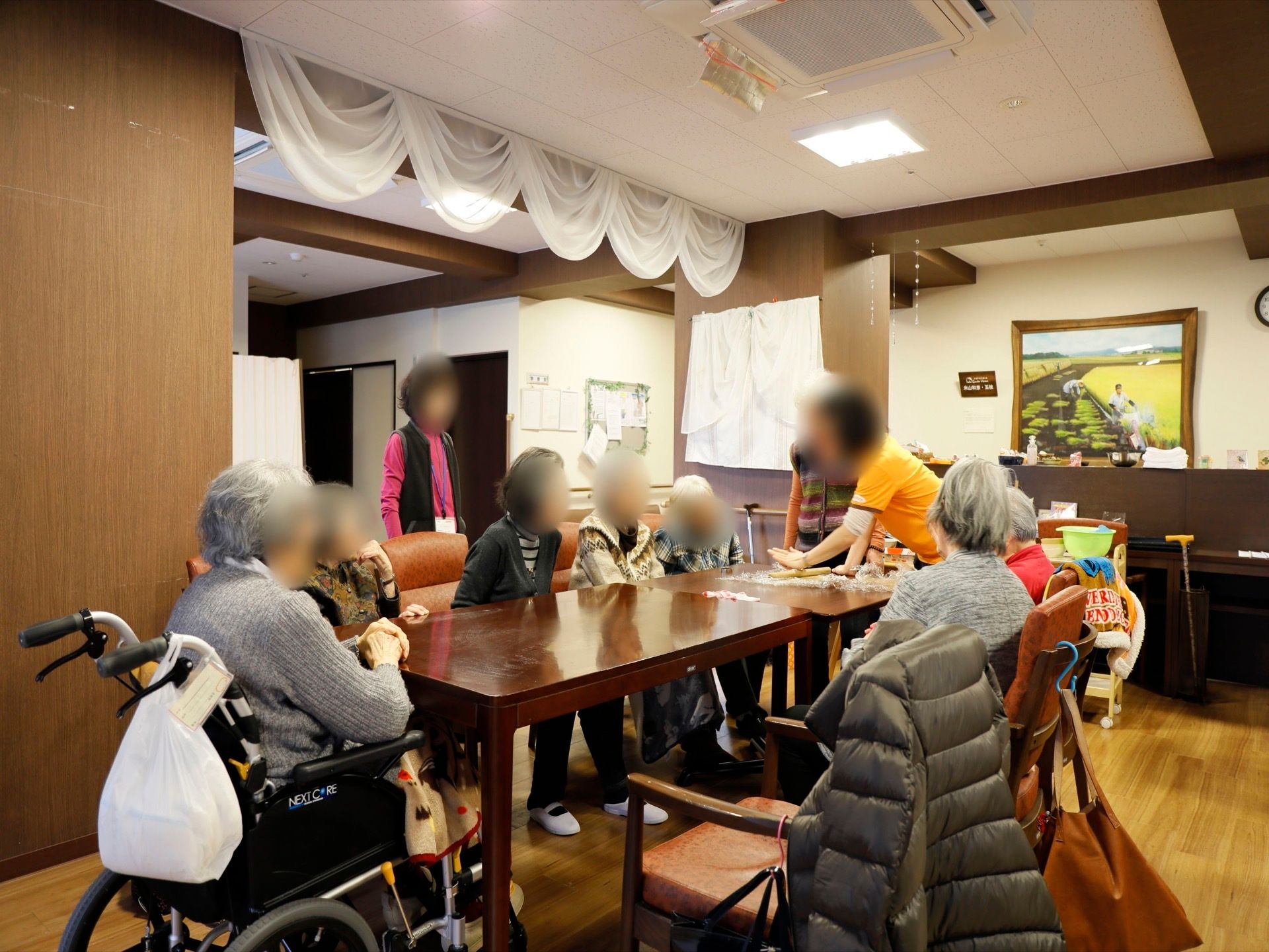 スタッフが指導の元、沢山の入居者様がテーブルを囲み活動を行っている様子。壁には絵が飾られ、天井には埋込式の空調が付いている。
