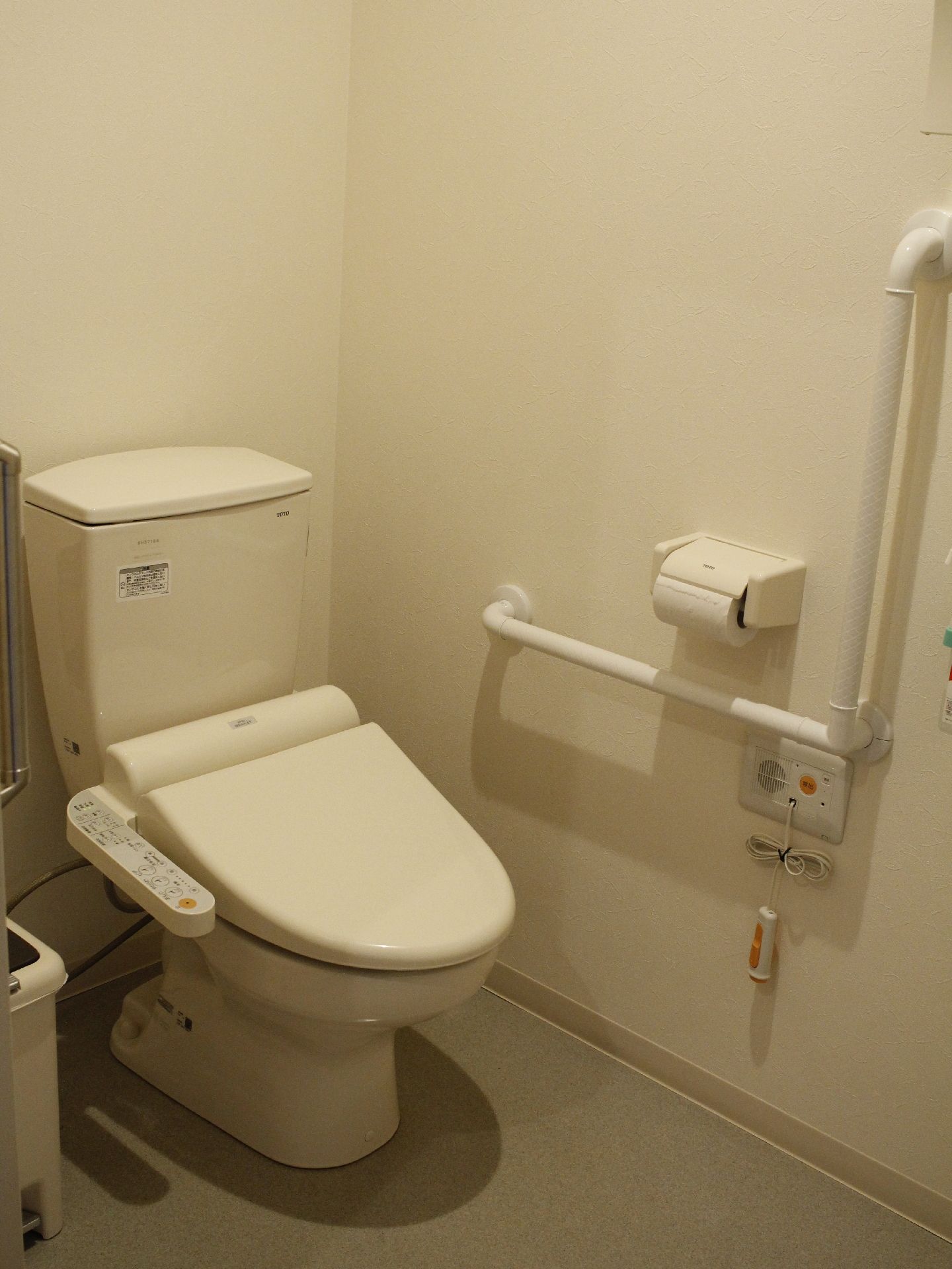 白色壁の内装のトイレスペースで、横の壁にはエル型の手すりとナースコール、トイレットペーパーが設置してある。
