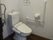 白色の壁に焦げ茶色の木目の床のトイレで、壁際にはエル型の手すりとタオル掛けが設置され、便器後ろのタンク上には水が出るようになっている。