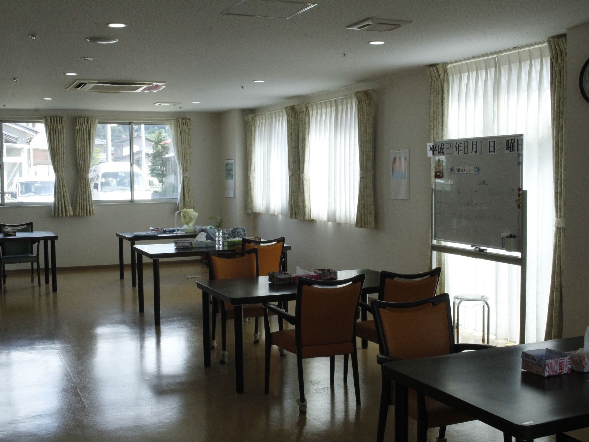 白色の天井と壁に、側面には、窓が並んでいる。4人掛けのテーブルと椅子が並んでいる。ボードが置いてあり、壁には時計がかけてある。