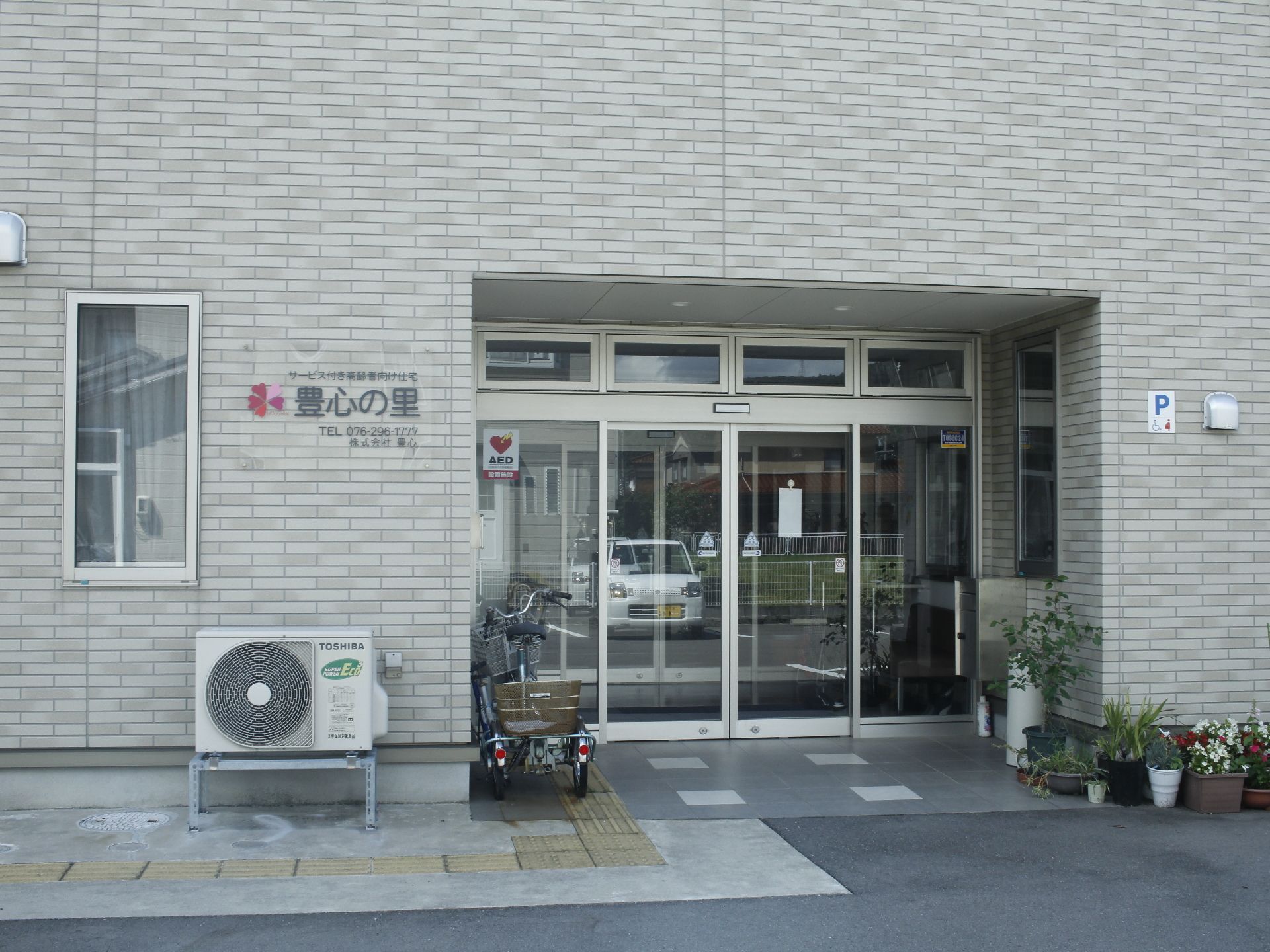 ガラス張りの二重ドアのエントランススペースで、玄関出入口傍には自転車が置かれ、建物の外壁傍には、植木鉢が並んでいる。