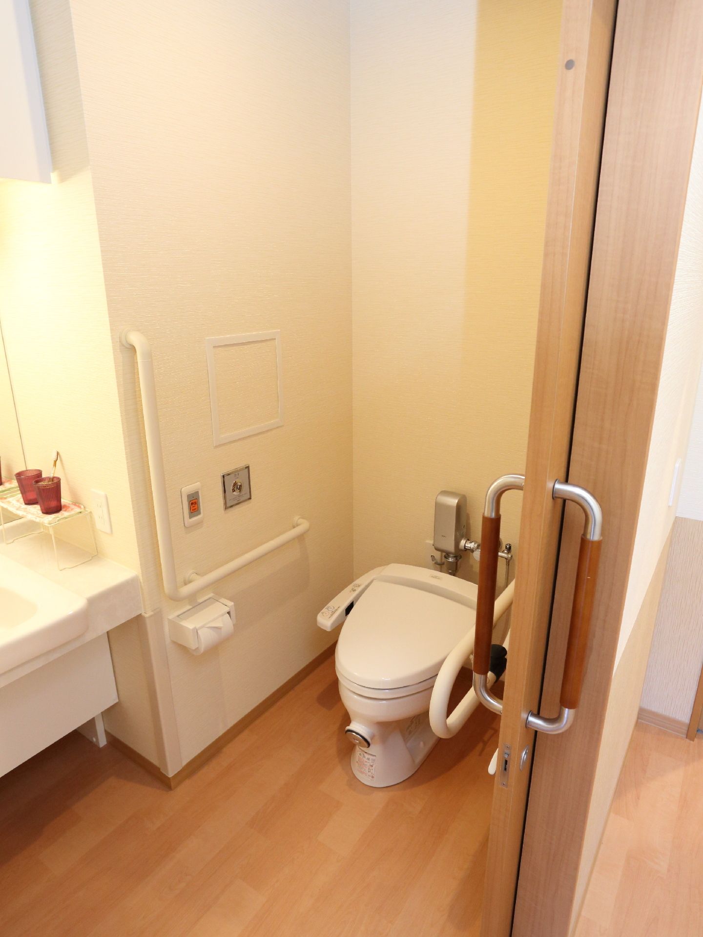 洗面器の横にコップが置いてあり、大面鏡が取り付けてある。白い手すりがトイレの壁についている。緊急時呼び出しのブザーがある。