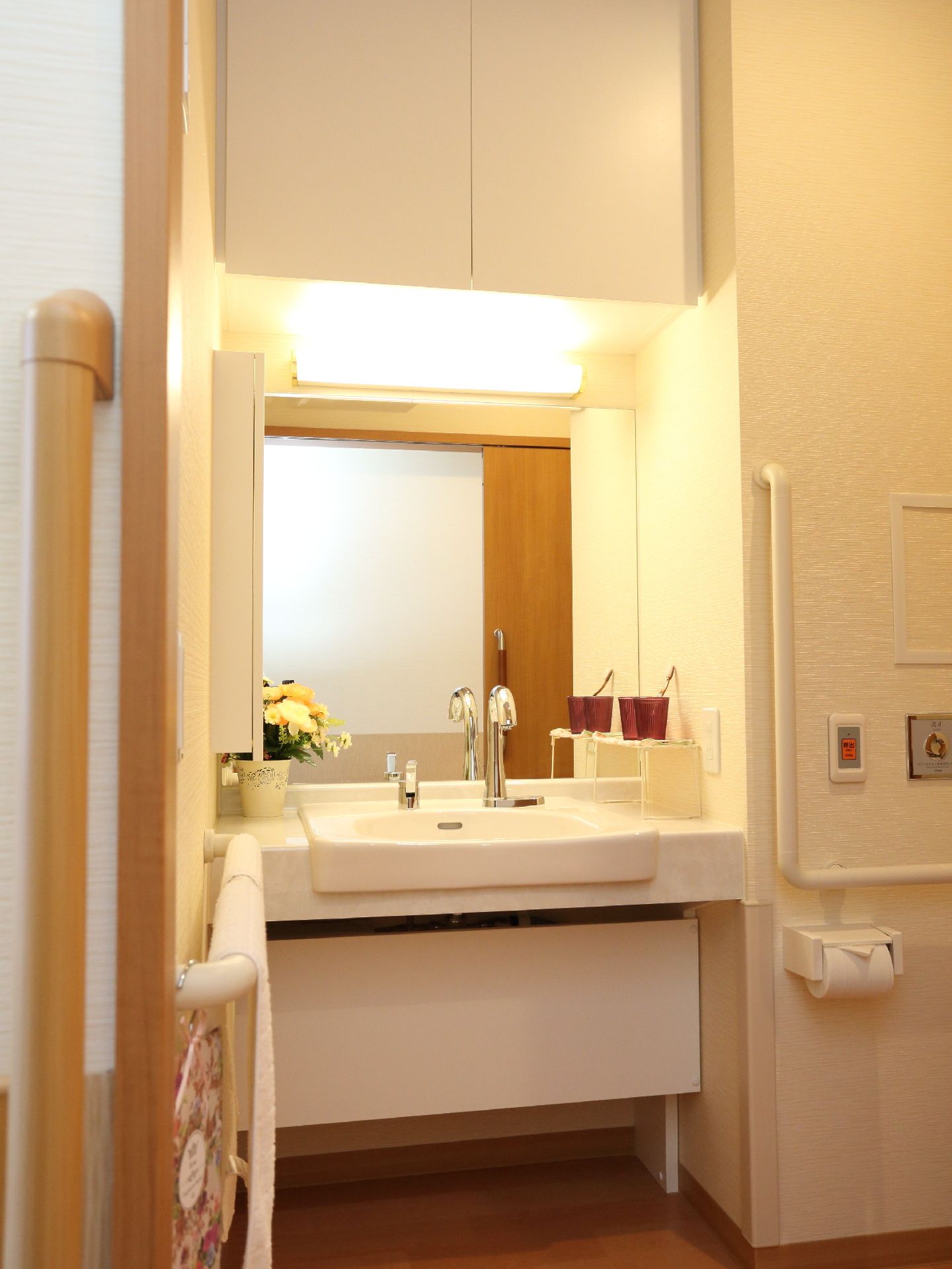 大面鏡が添えてある洗面台で、花が活けてある。収納棚がついている。手すりがトイレの壁の両側についている。
