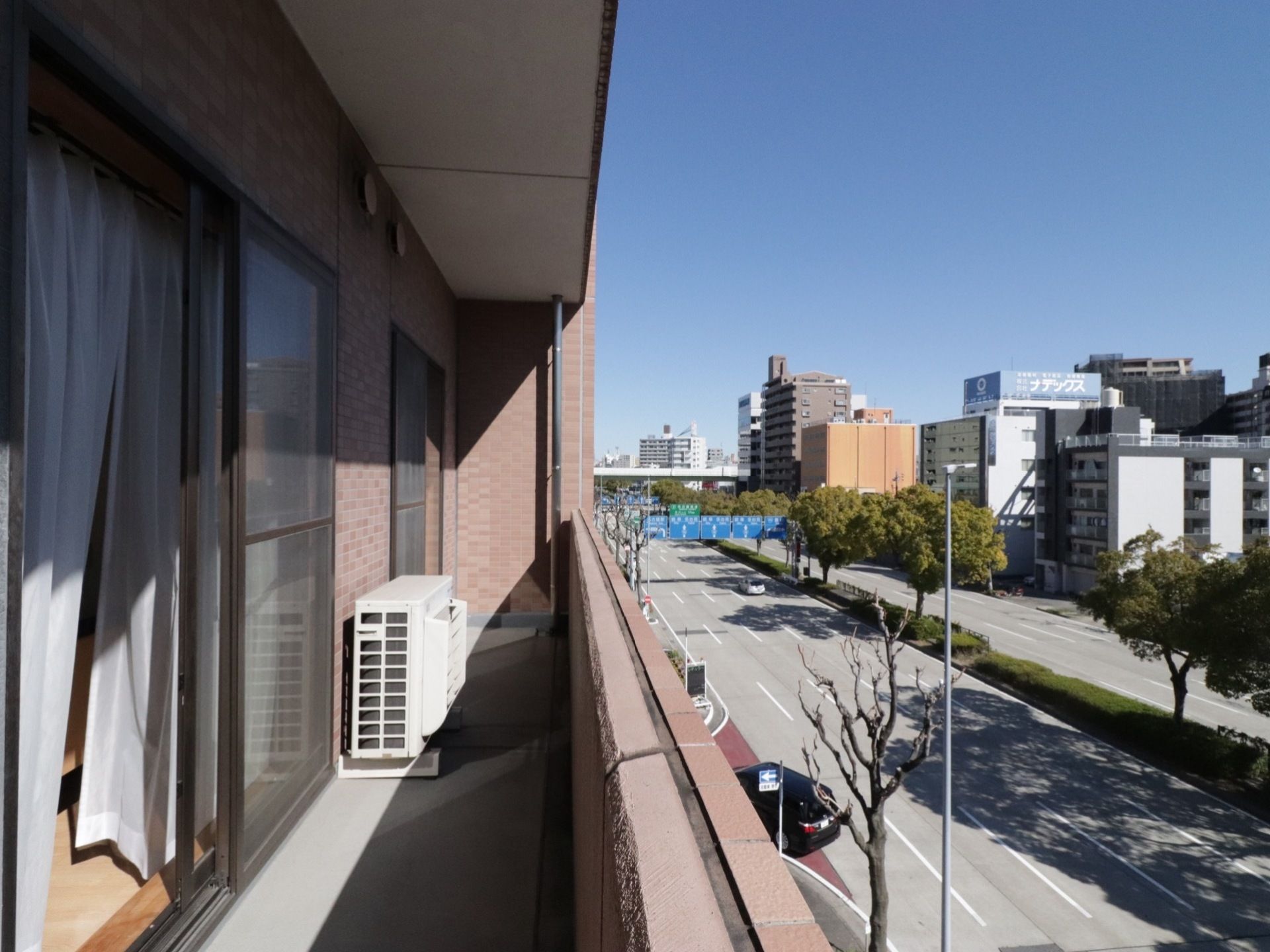 バルコニーには室外機のみがあり、広々としている。近くに高い建物が少なく、窓からは開放的に青空と町並みを一望することができる。