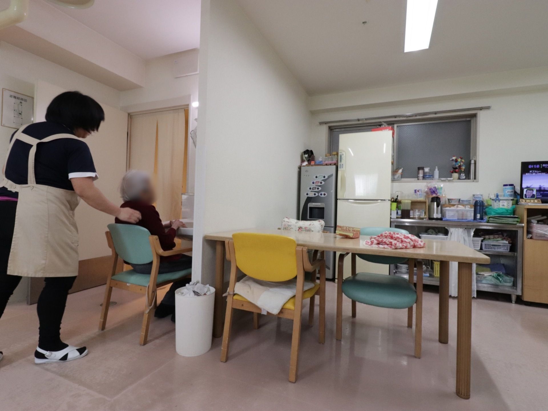 ケアスタッフがマンツーマンで入居者様のサポートをしている。部屋にはテーブル、椅子、冷蔵庫、給茶機、テレビがある。