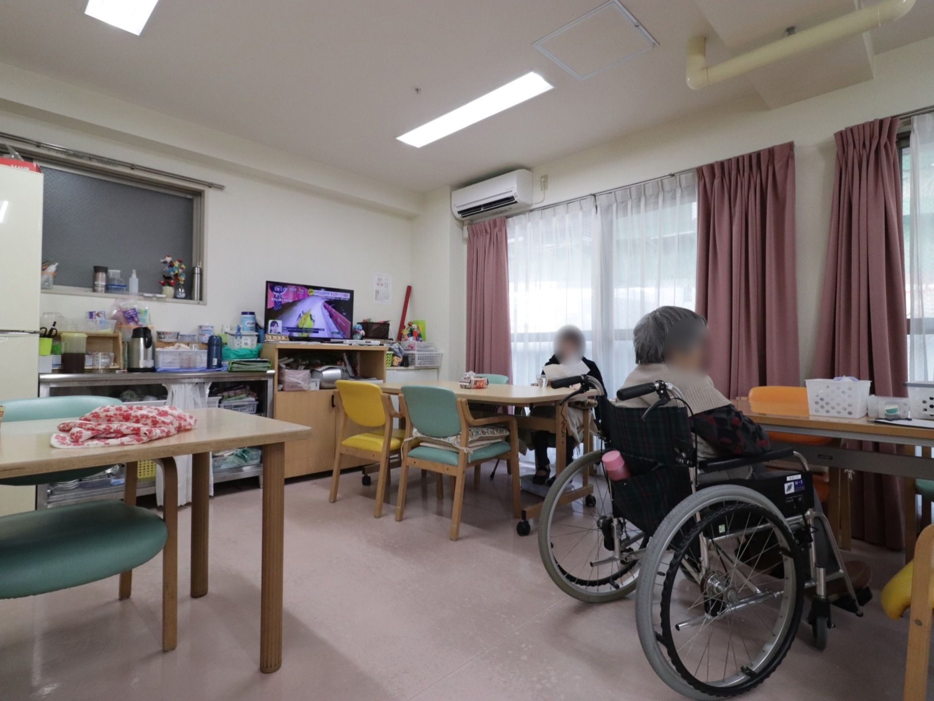 明るく広いスペースにテーブルと椅子、冷蔵庫、給茶機、テレビが設置されている。入居者様がそれぞれに作業をされている。