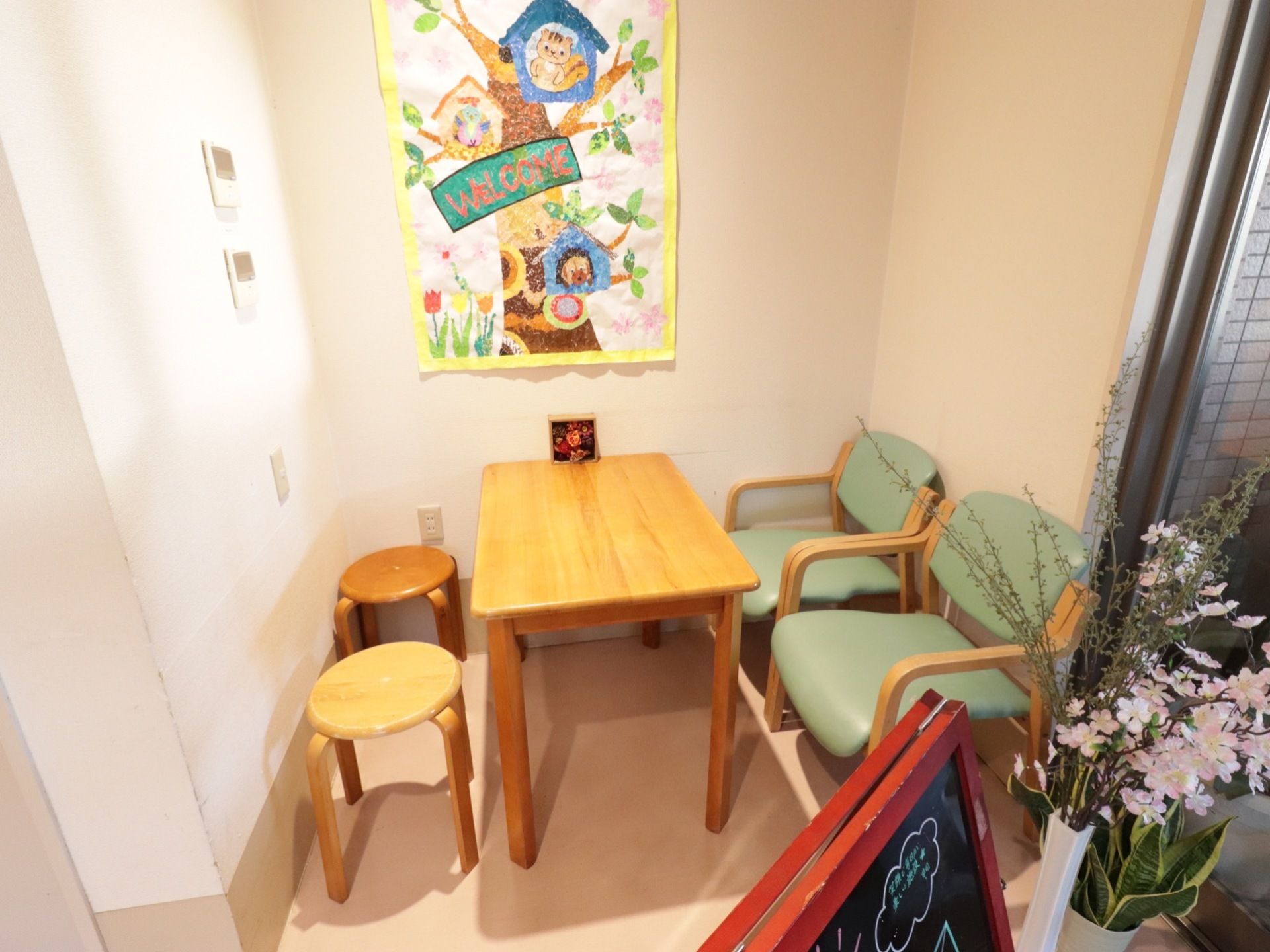 テーブルと椅子が４脚あり明るい空間。花瓶には花が飾られ、壁には手製のウェルカムポスターが貼られている。