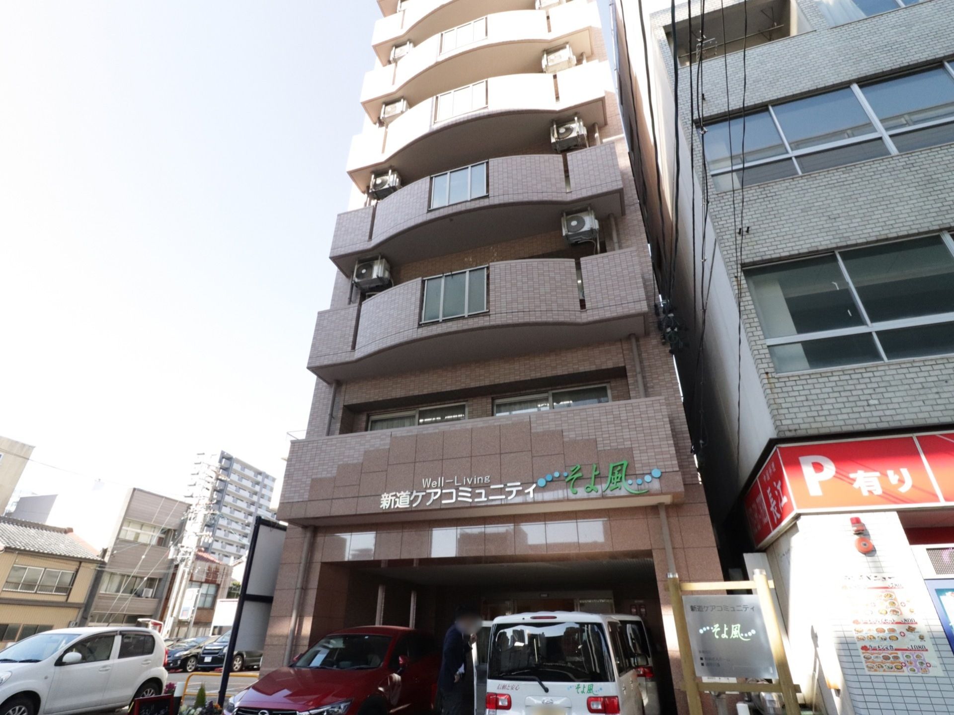 施設は6階以上からなる建物である。1階部分には車が2台止められている。建物の隣は駐車スペースになっている。