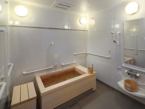 白い壁の浴室に檜の浴槽や移乗台が置かれており、壁には横型や縦型の手すりが設置されている。大きな鏡の前のシャワーコーナーにも手すりがある。
