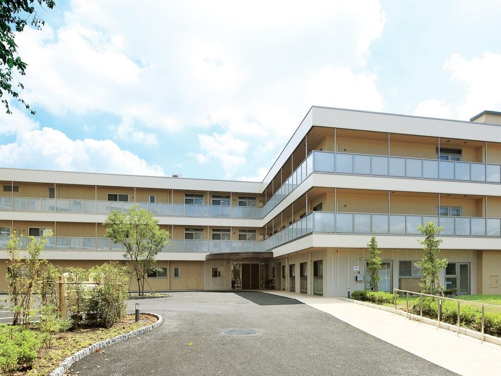 ベージュの外壁と広いベランダが目を引く、緑に囲まれた３階建ての入居施設。建物入口まで道路が整備されており、歩道や手すりも設置されている。