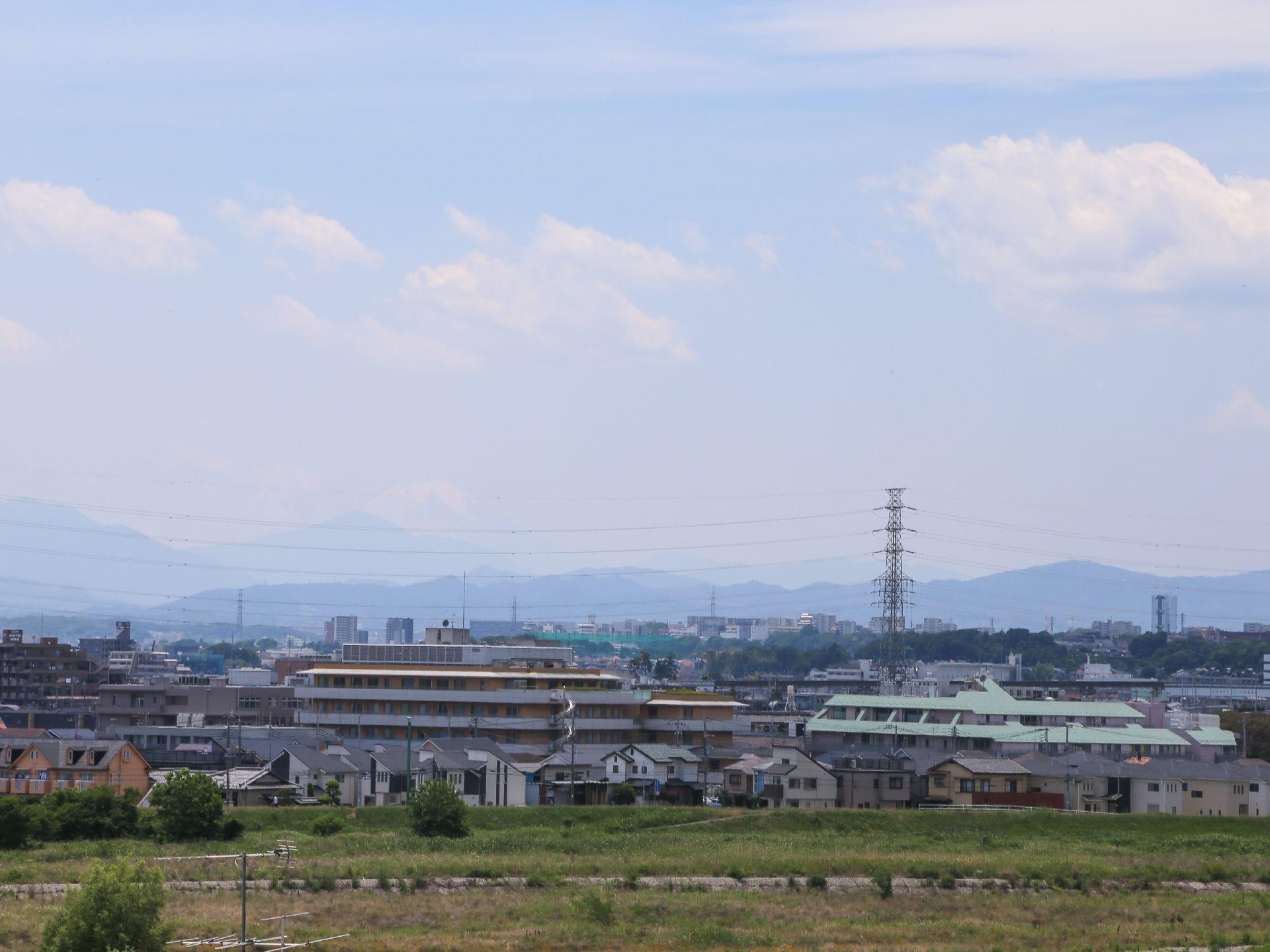 施設周囲の景色の写真である。遠くに山々を望み、その手前に鉄塔と高圧電線が渡っている。施設前は空き地が広がっている。