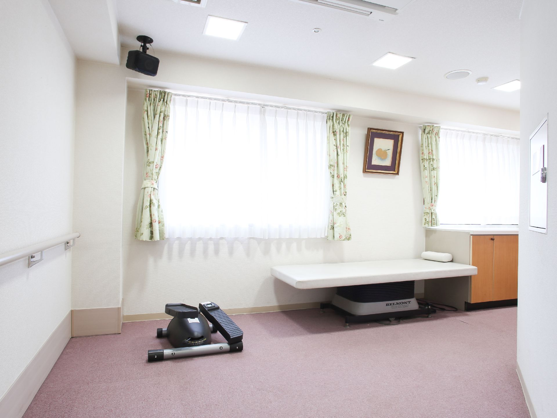 大きな窓が２か所にあり、レースのカーテンが閉められている。窓の前に診察用のベッドがあり、その横にトレーニング器具がある。
