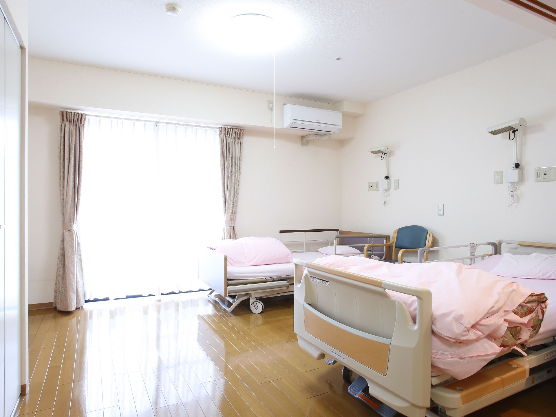 ２人用の居室で、壁際に介護用ベッドが２台ある。ベランダへ出られる大きな窓にカーテンがかけられている。