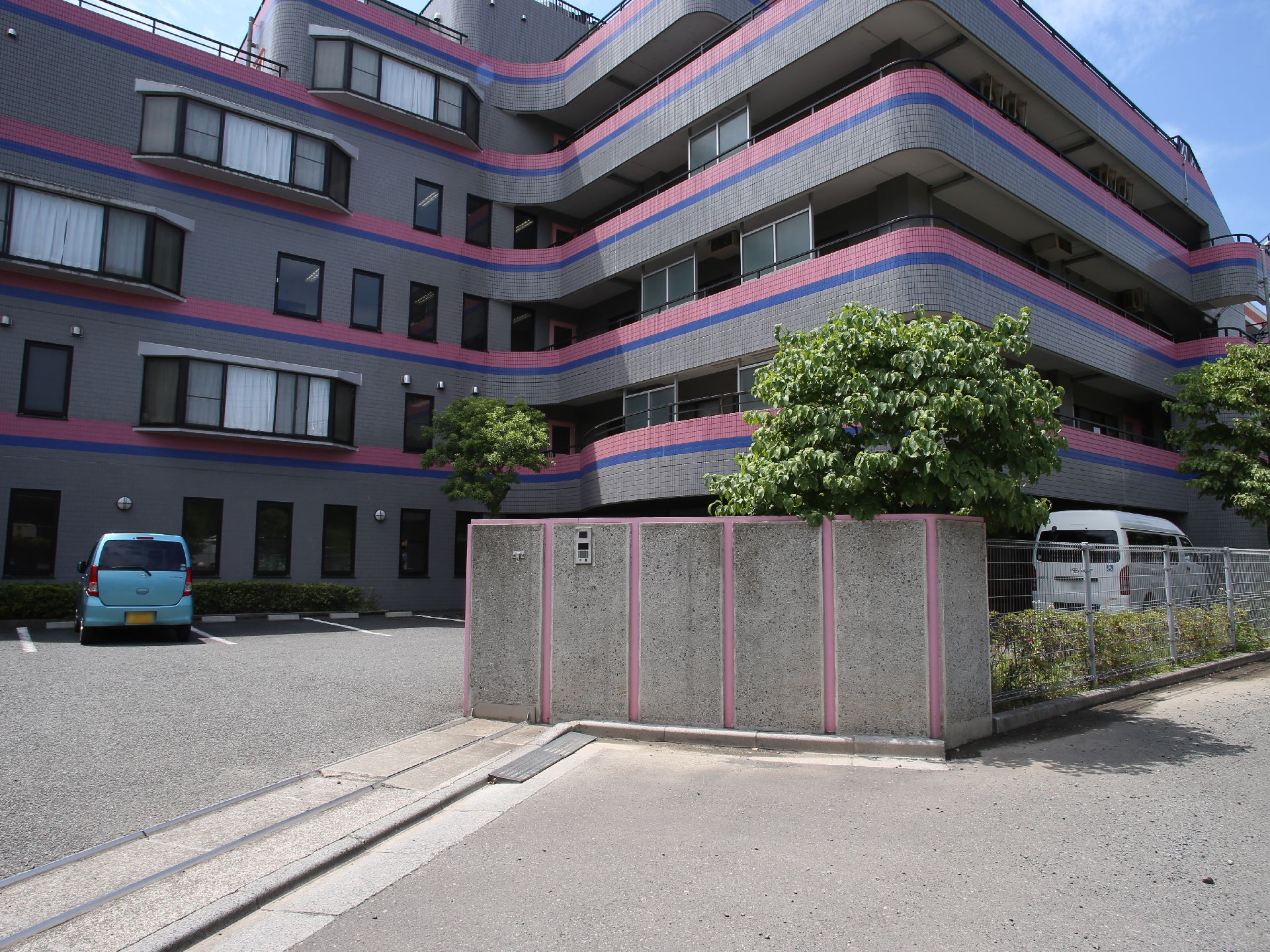 グレーの外壁に、ピンクと青のラインが入った建物である。敷地入口にはコンクリート製の門があり、インターホンが取りつけられている。