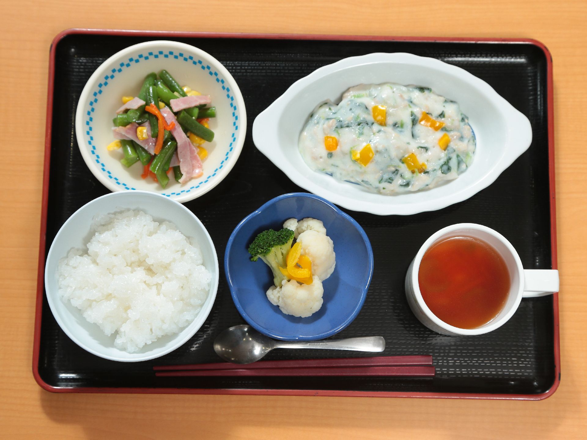黒い和風のトレイに赤い縁取りがされている。左側にごはんとゆで野菜、小鉢があり、右側にスープとメイン料理が乗っている。