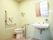 白いタイル張りの個室トイレで、左奥に洋式便座がある。便座の両脇には固定式の手すりがあり、壁にトイレットペーパーがある。