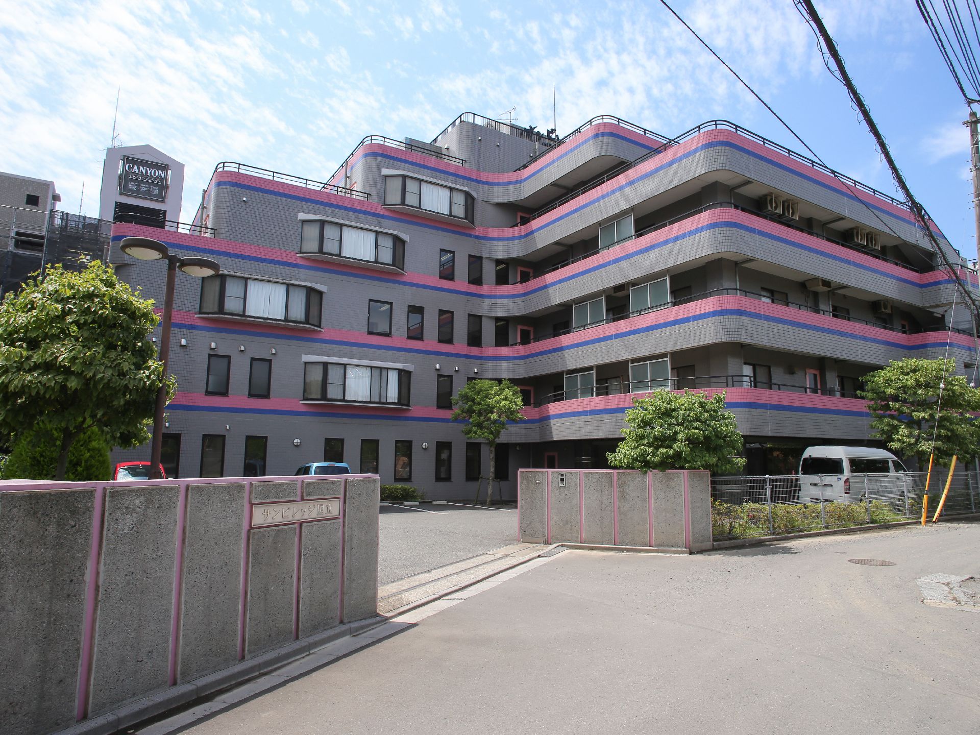 ６階まである建物で、濃いグレーの外壁にピンクと青のラインが入っている。敷地入口は門があり、施設に沿って植栽が植えられている。