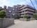 ６階まである建物で、濃いグレーの外壁にピンクと青のラインが入っている。敷地入口は門があり、施設に沿って植栽が植えられている。