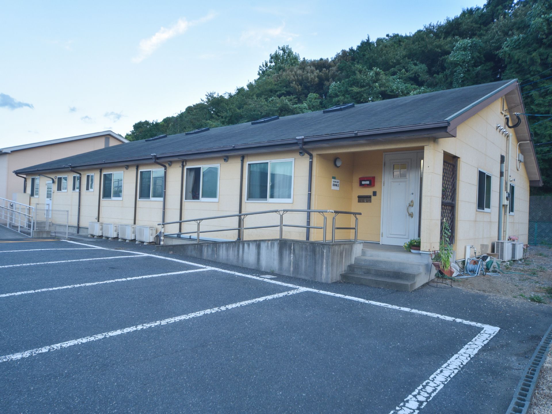 建物の前に何台も停められる駐車場がある。玄関の前には階段とスロープがある。建物の後ろには豊かな緑がある。