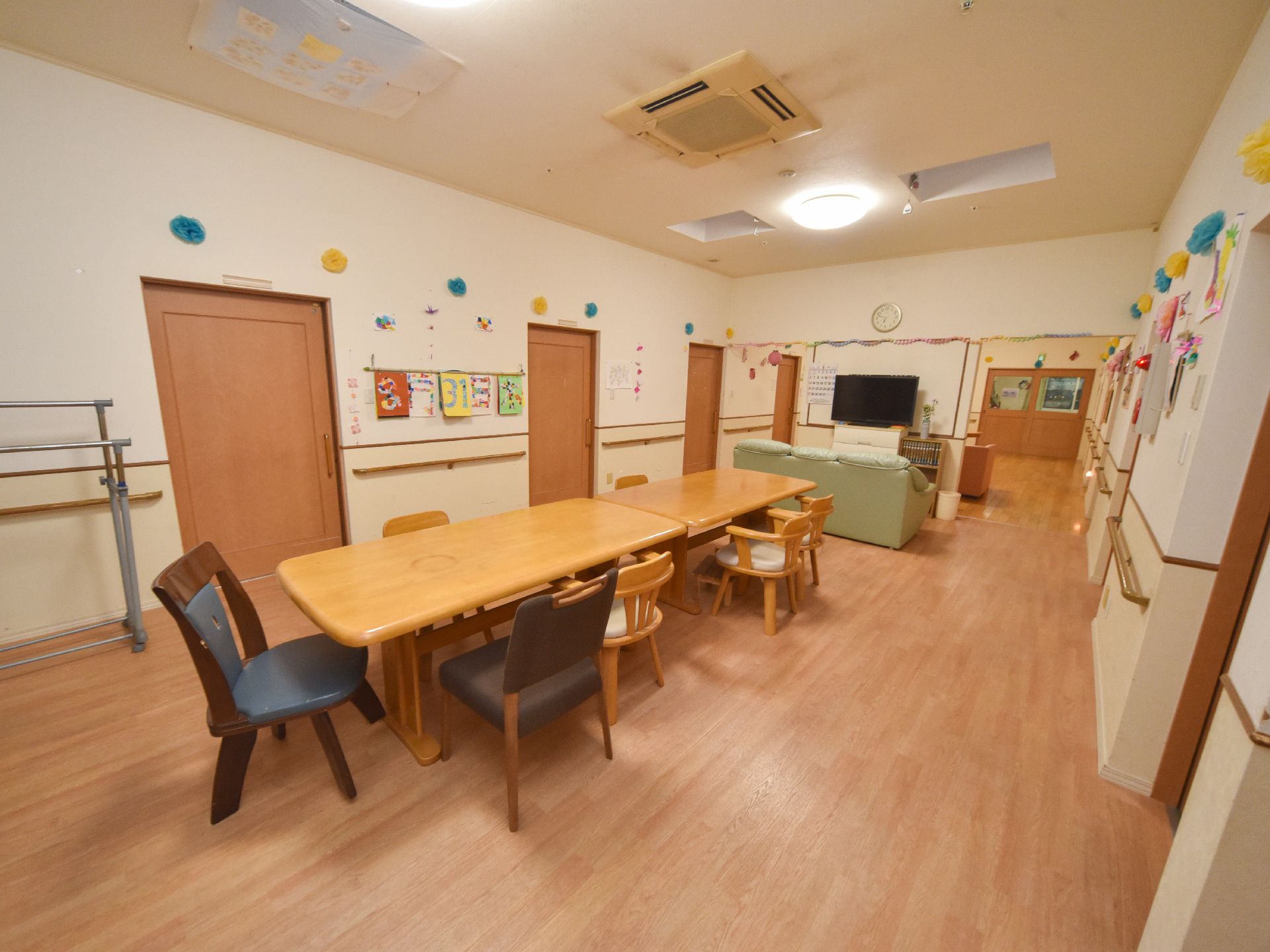 壁には手作りのカレンダーと飾りが貼られている。扉は幾つもあり全て引戸である。手摺もある。机と椅子、ソファーにテレビが置かれている。