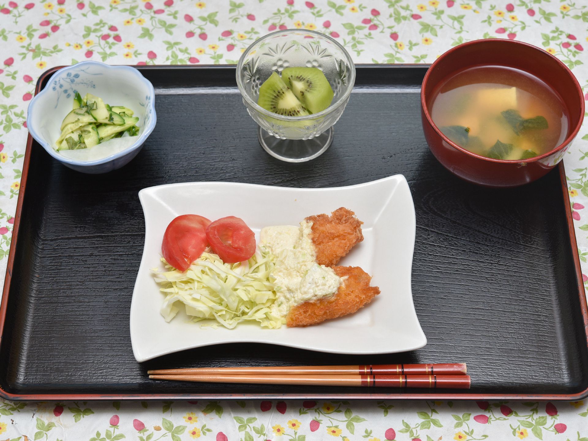 花柄のテーブルクロスの上に置かれた木製のお盆。その上には、メインのおかずとサラダ、漬物、味噌汁、果物がのっている。