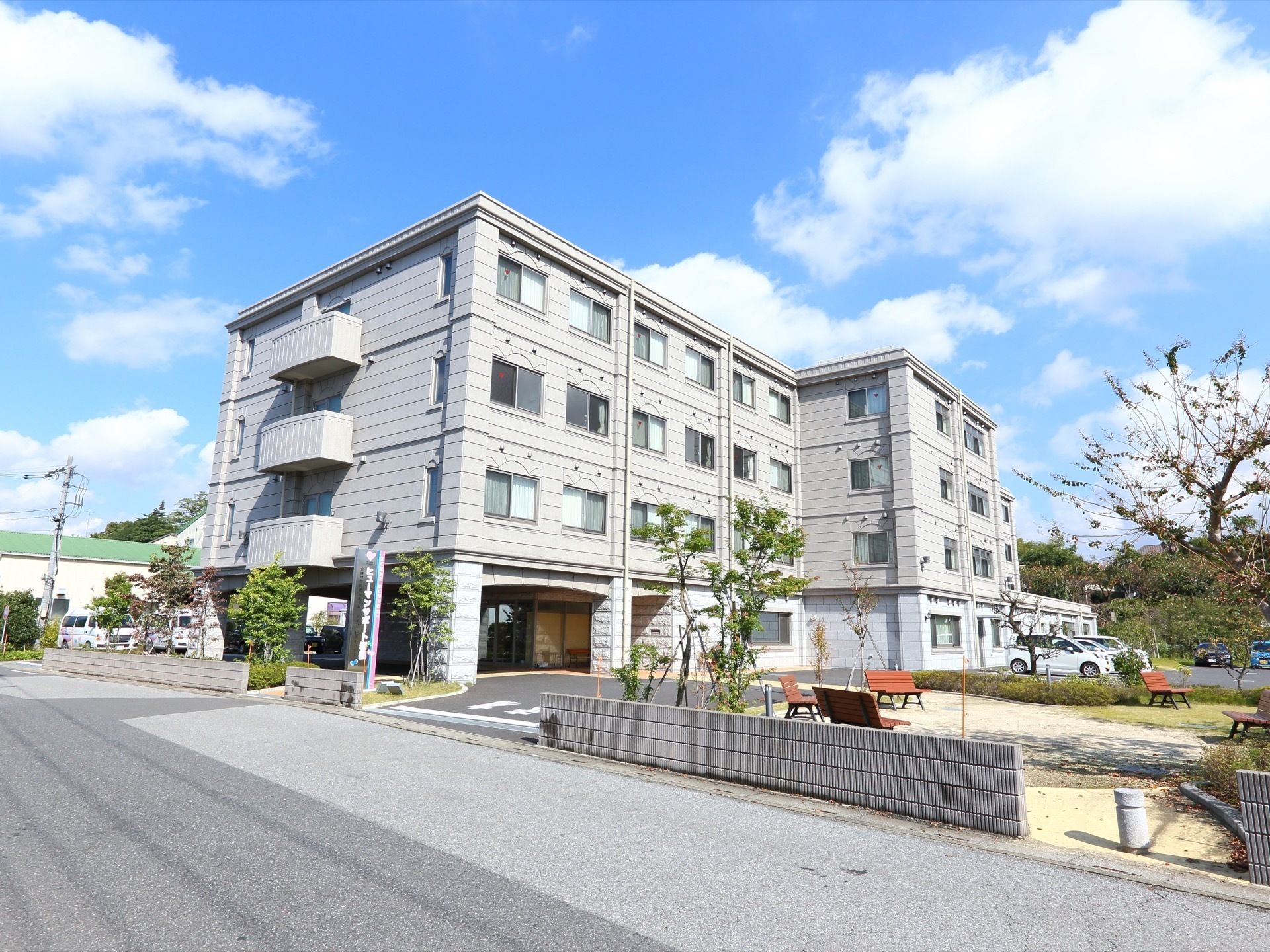 青空の快晴の下、４階建ての施設外観が写っている。周辺にはたくさんの植物が生えており、車が何台か停まっている。