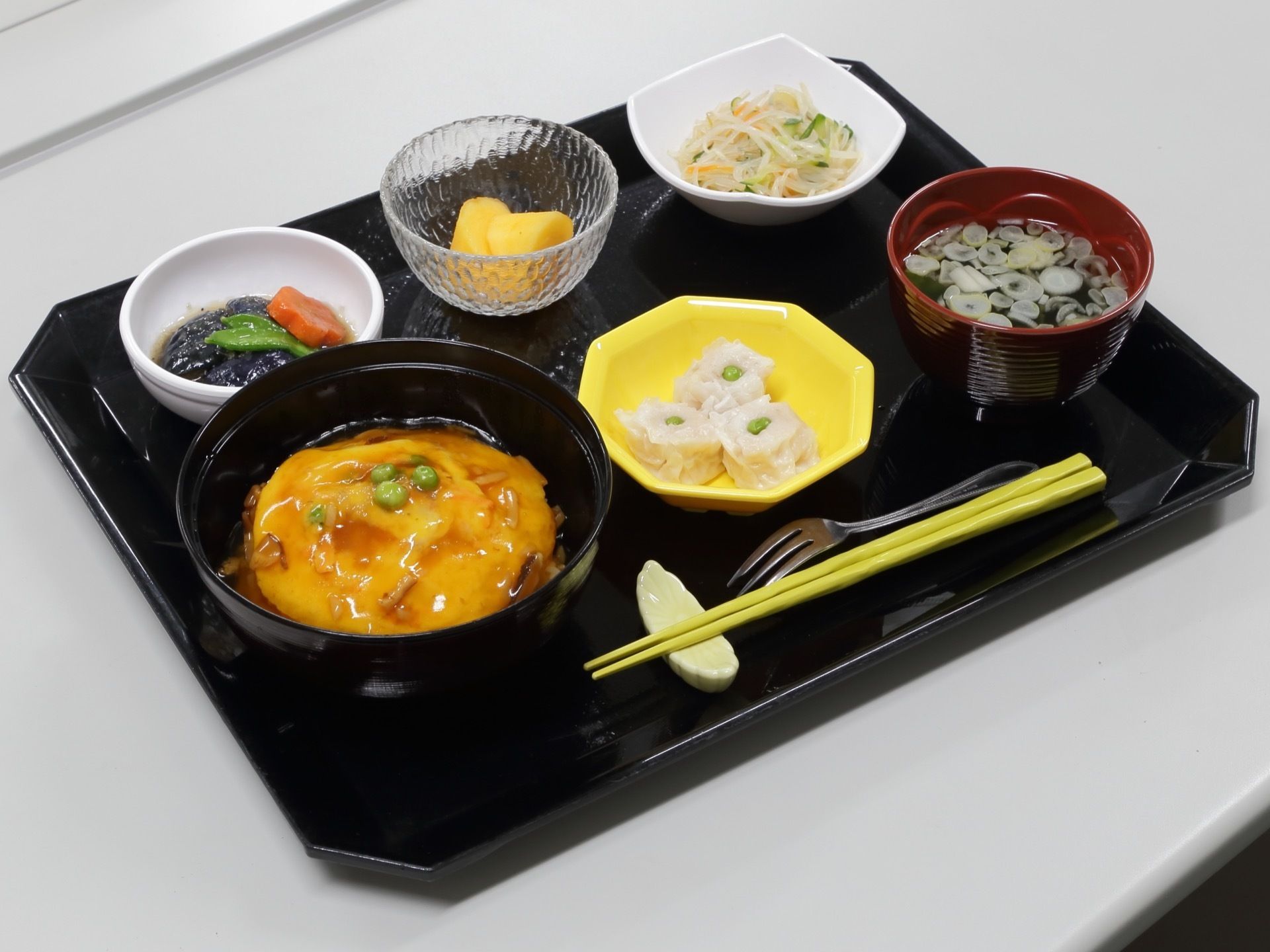 黒いお盆の上には様々な食材がのせられている。手前には箸置きにのった黄色の箸やフォークが添えられている。