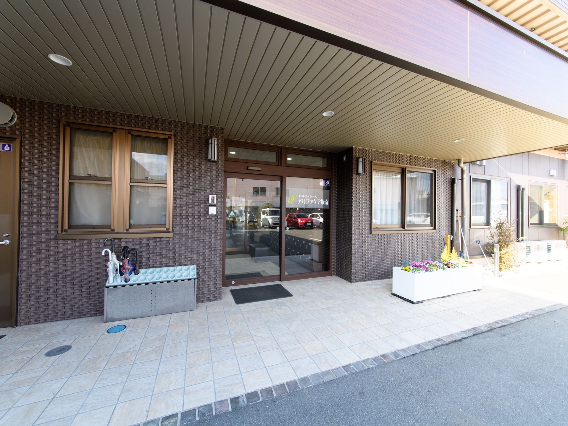 自動ドアでバリアフリーになっている。ドアに施設名が書かれている。左側に傘立てが置かれ、右側に花壇がある。玄関の両側に窓がある。