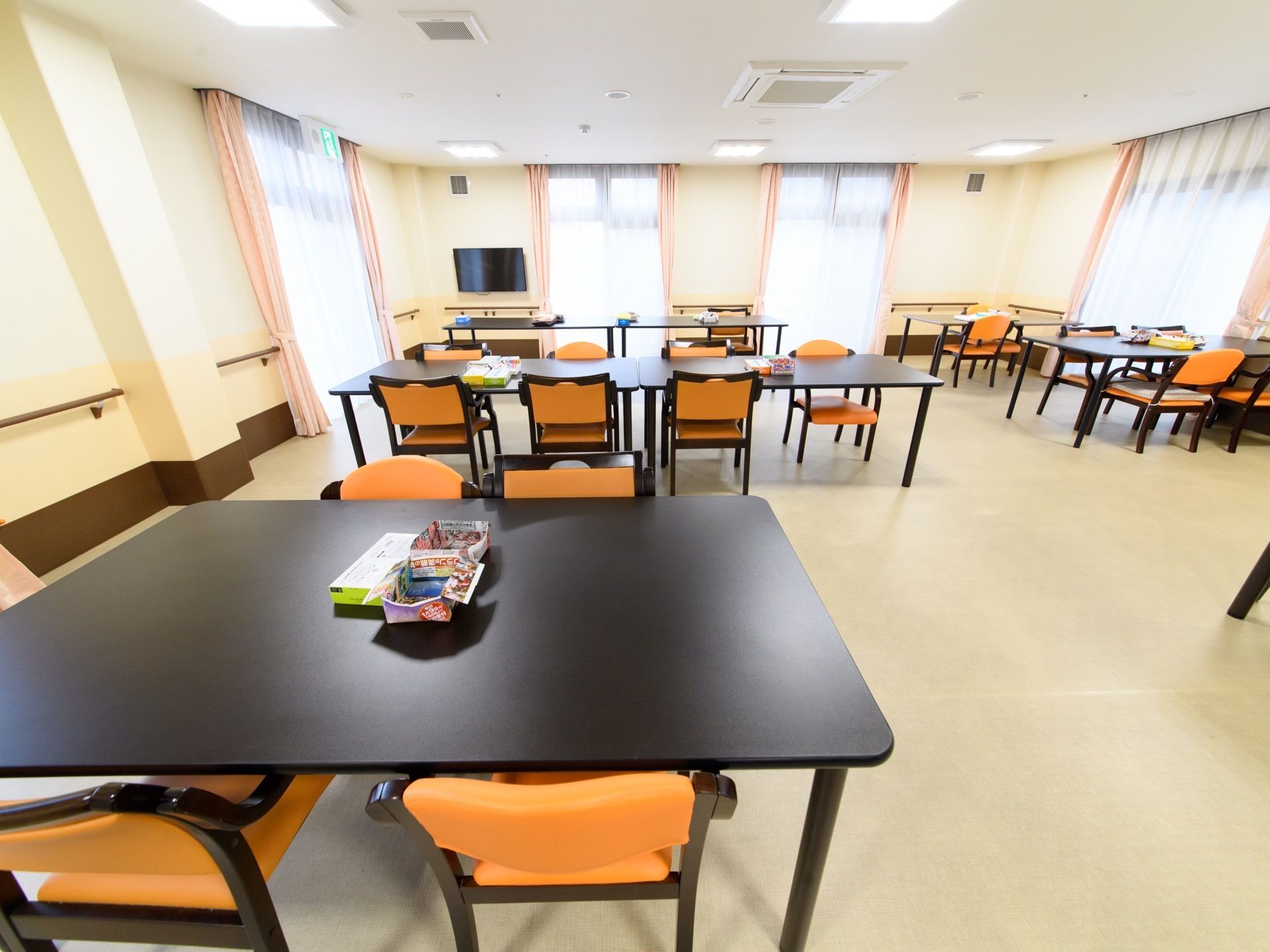 テーブルと椅子がたくさん並んでいる。窓があり、白色のレースとピンク色のカーテンがついている。テレビが設置されている。
