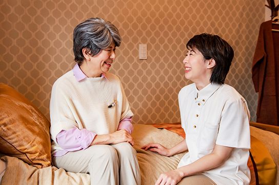 カーディガンを着た高齢女性が高齢男性と一緒にソファに座り、二人とも微笑んでいる。