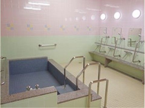 複数の照明が設置されている明るい大浴場。浴室の壁には手すりがあり、洗い場に３人分のシャワーや鏡が取り付けられている。