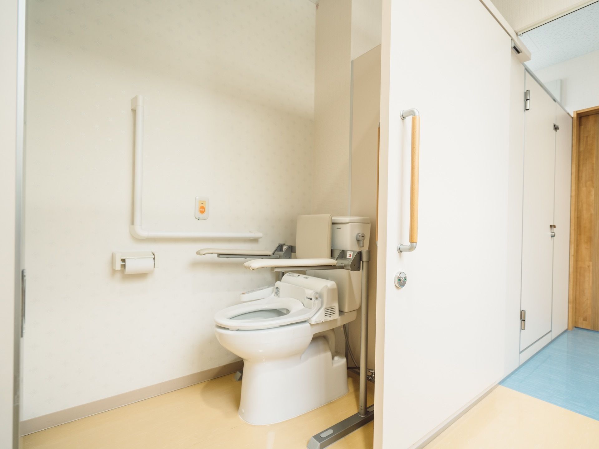 白い壁とドアの個室トイレで、背もたれのついた洋式便座が1台設置されている。便座の左右両側に可動式のアームレストがある。