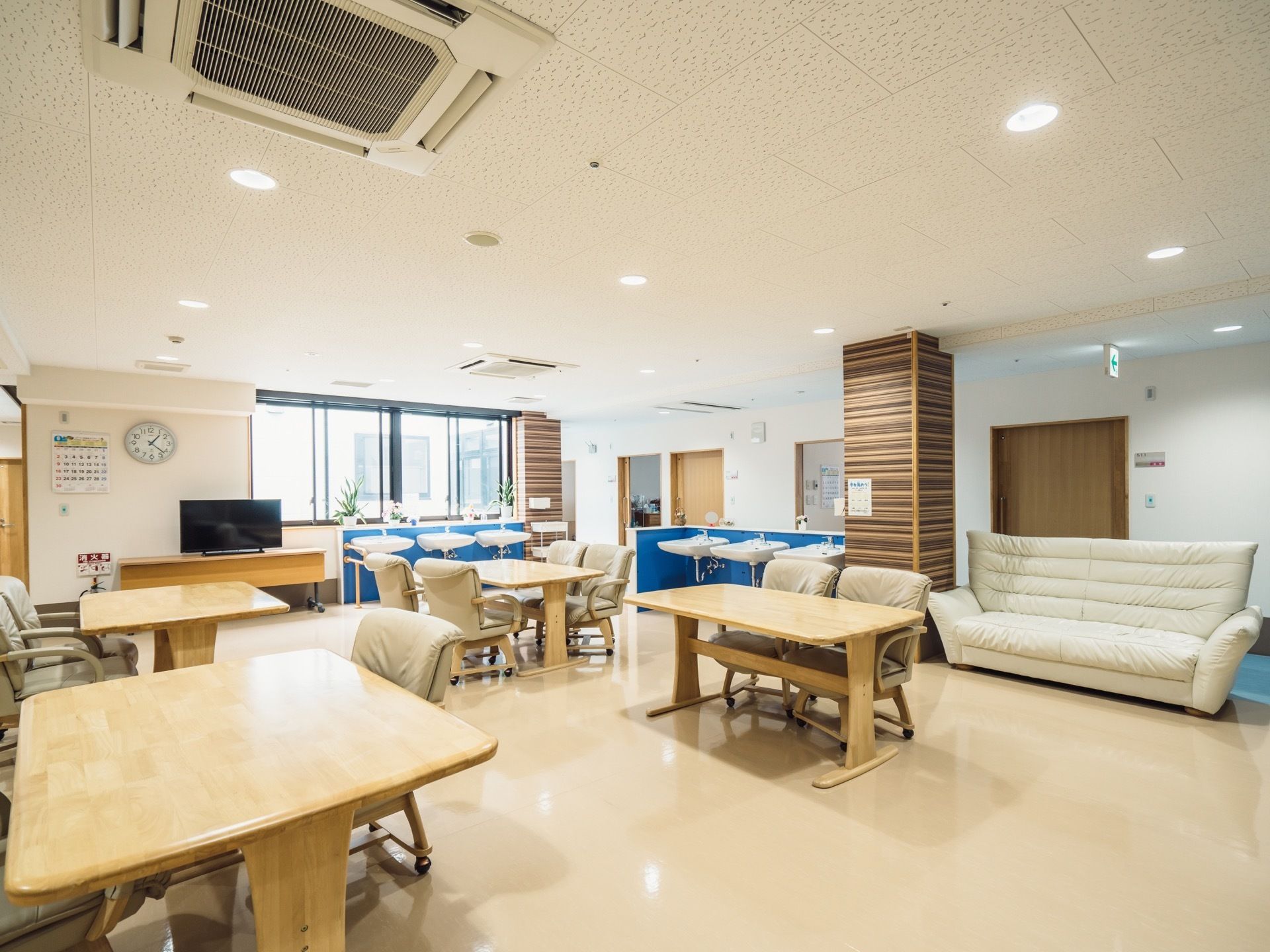 窓側に黒いテレビを置いた木製の台がある。テレビの上の壁に丸型の時計とカレンダーが取り付けられている。