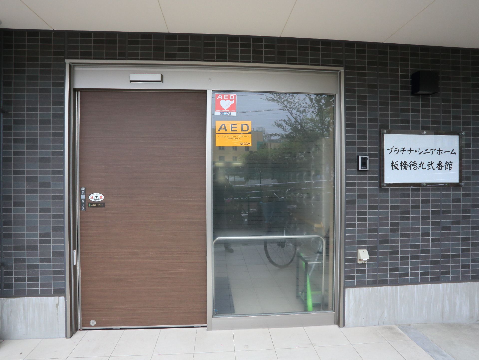 黒が基調のレンガ調の外壁には施設名の看板があり、大きな文字でわかりやすい。ブラウンの木目調の自動ドアになっている。