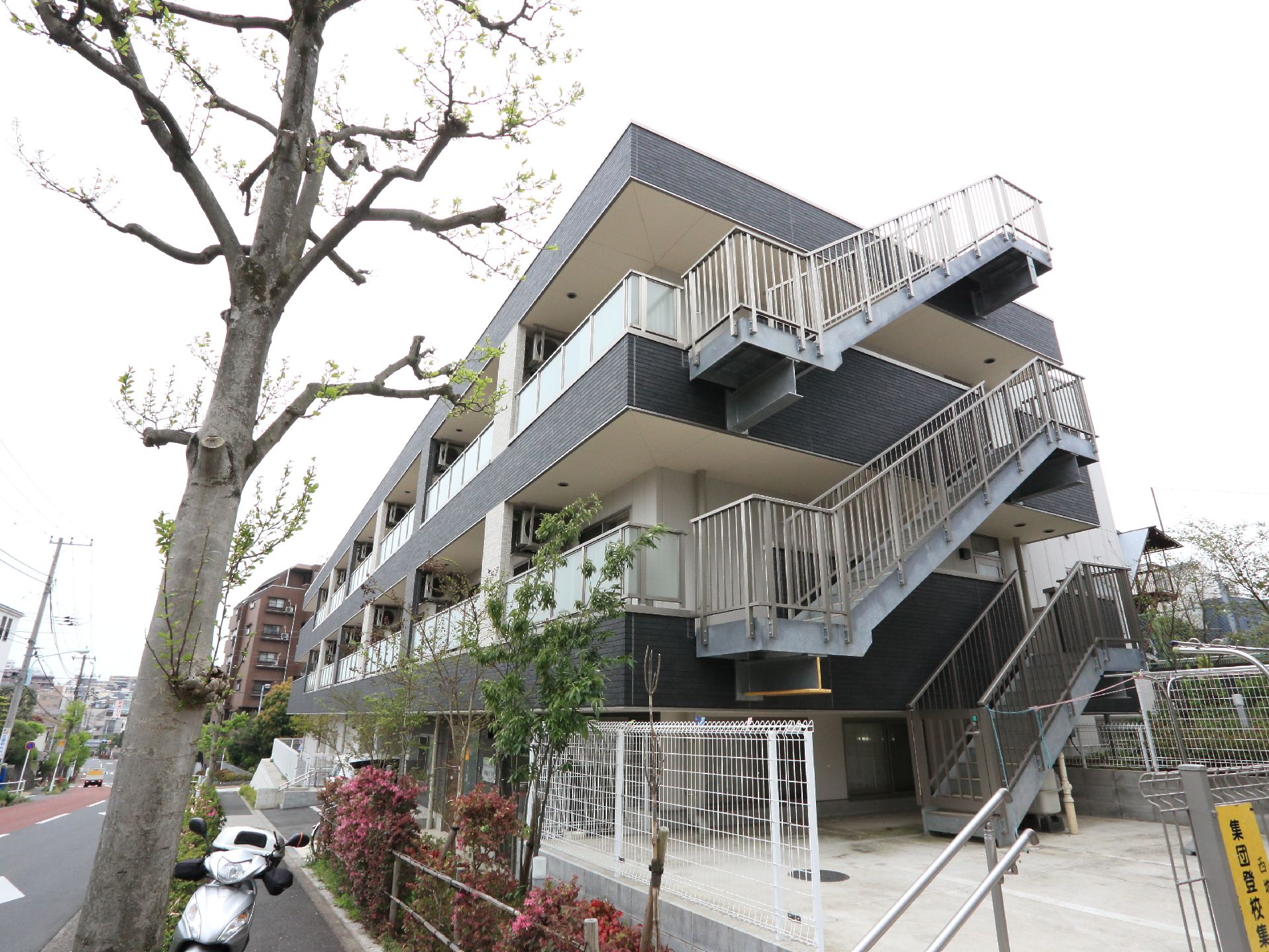 黒を基調とした外壁の建物で、3階建てになっている。非常階段があり、白い柵も見えている。建物の周りには木々も見える。