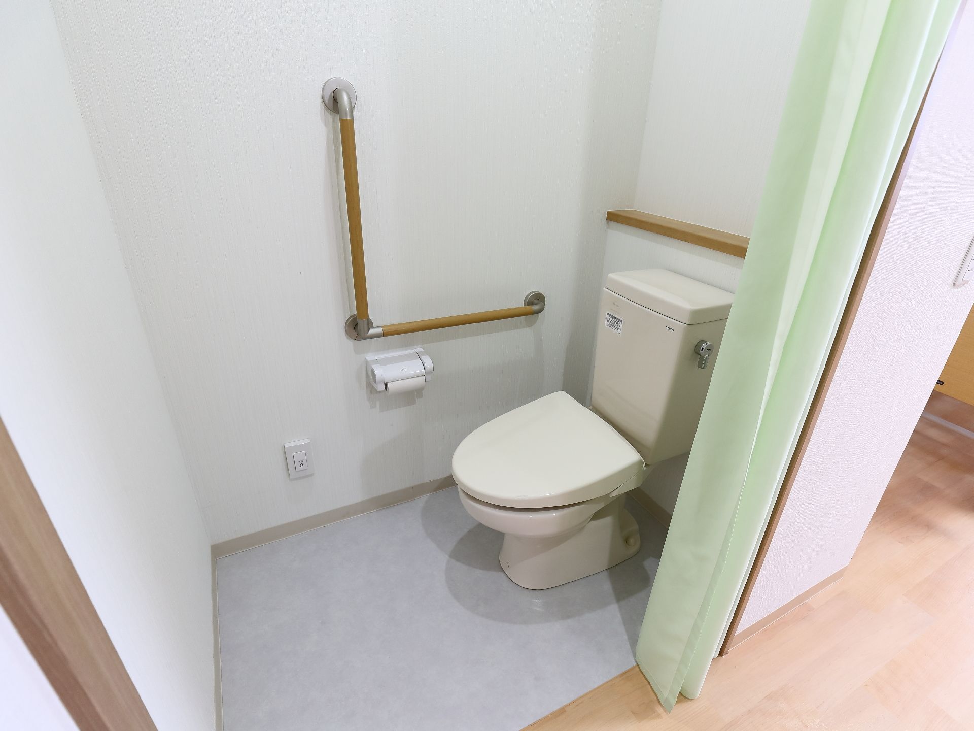 居室備え付けのトイレである。共有トイレに比べシンプルな機能で、仕切りにはドアではなくライトグリーンのカーテンが使われている。