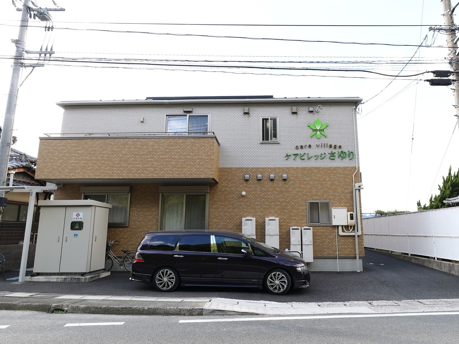 22名の定員の施設が鉄筋２階建てのコンパクトな建物に収まっている。表には施設所有及びスタッフ用の車両が停まっている。