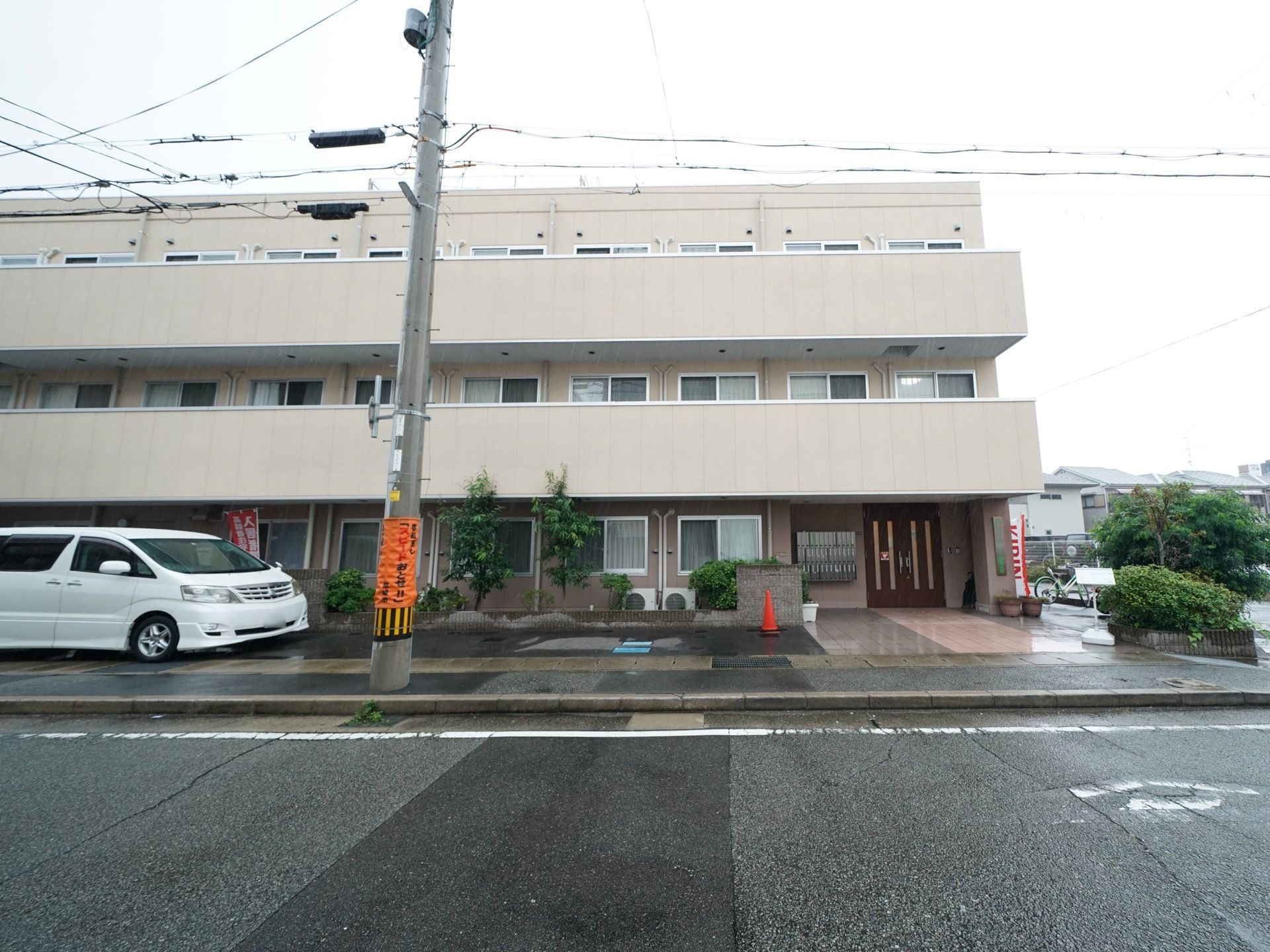 ２階と３階にベランダが設置された白い３階建ての建物がある。建物の前の敷地に白い大きな車が停められている。