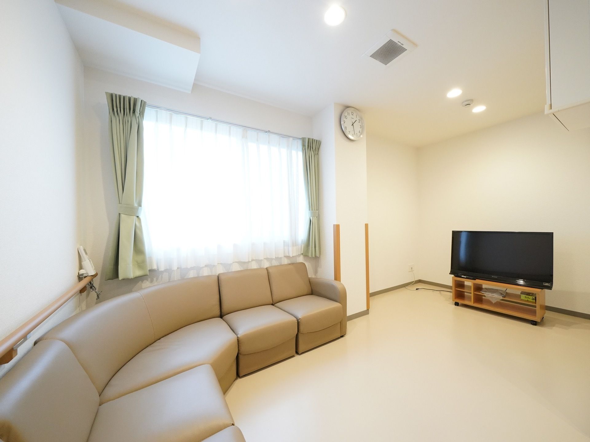 黒い薄型テレビが置いてあり、壁に時計が掛けられている。ベージュ色の大きなソファーの背後に腰高窓が設置されている。