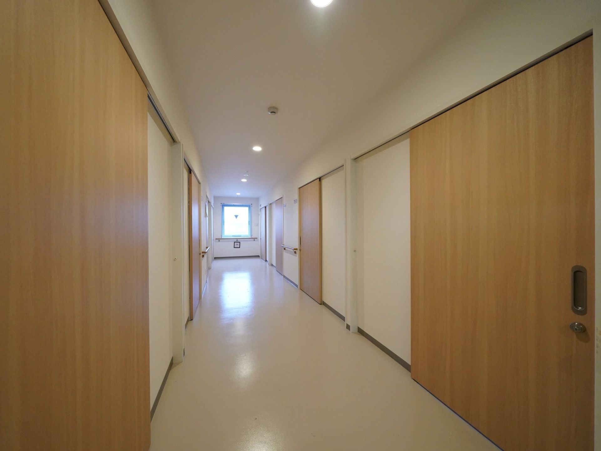 清掃が行き届いた長い通路の両側に、各居室へ入るスライドドアがある。通路の突き当りに窓が設置され、光が差し込んでいる。