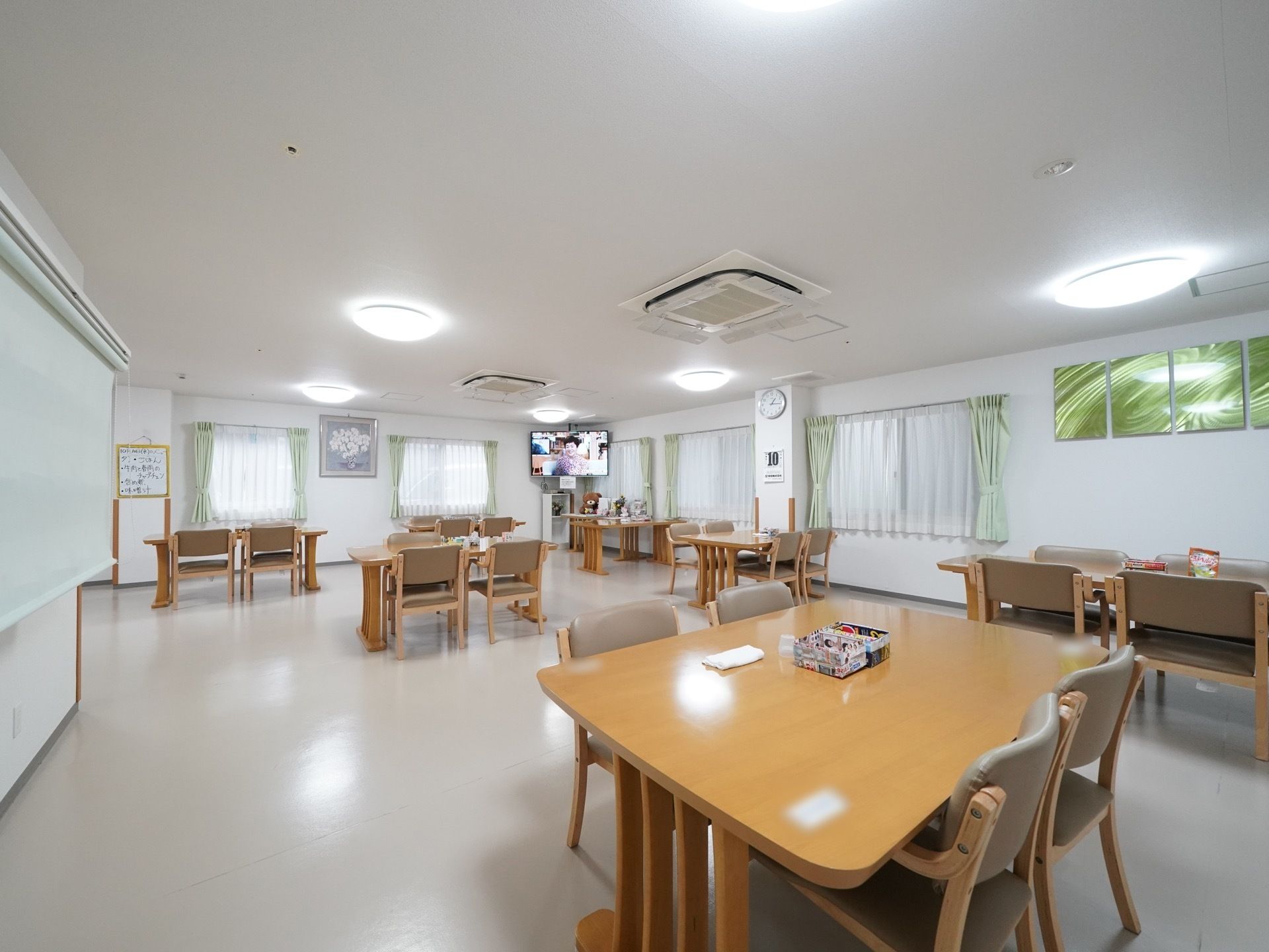 広々とした室内に、ゆとりをもって数組のダイニングセットが配置されている。黄緑色のカーテンが掛けられた腰高窓が複数個所にある。