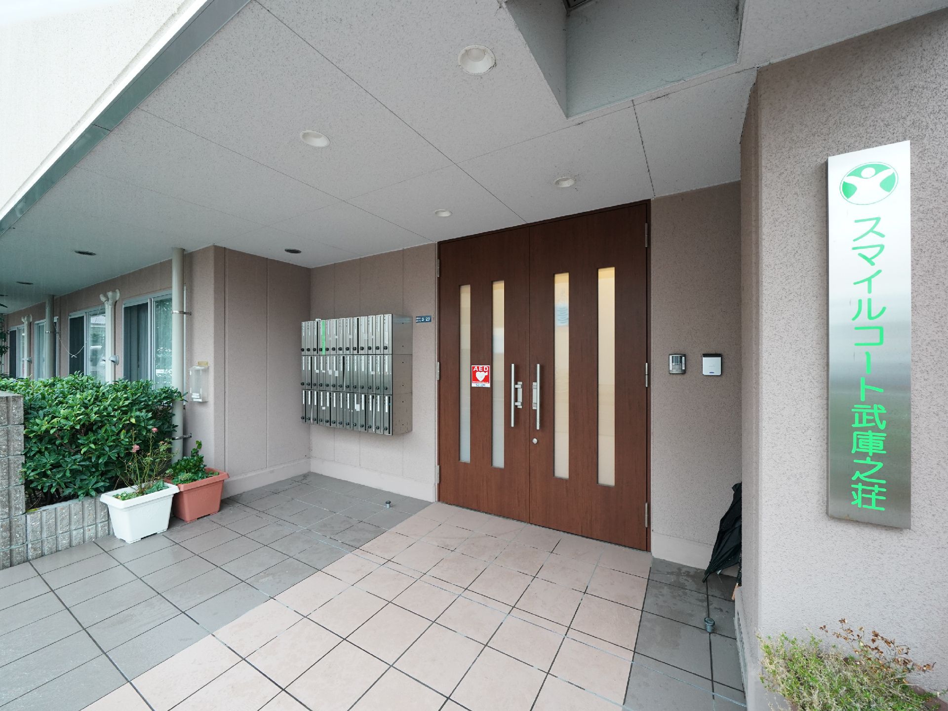 ベージュの壁にブラウンの両開きのドアが配され、右側に看板のかかった玄関