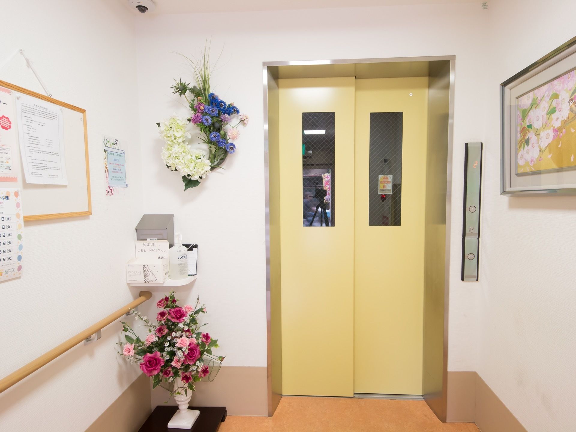 玄関脇には綺麗な花を飾っている。壁には手すり、天井には防犯カメラを設置している。ボタンは車椅子でも押せる高さになっている。